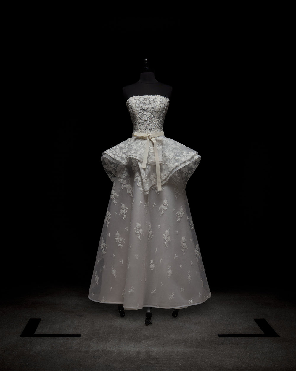 The Viviana Dress by SVT DESIGNS is a strapless white gown on display, featuring intricate lace detailing, a fitted bodice with a flared peplum, and a long flowing skirt adorned with floral patterns, complete with a waist-accentuating bow.