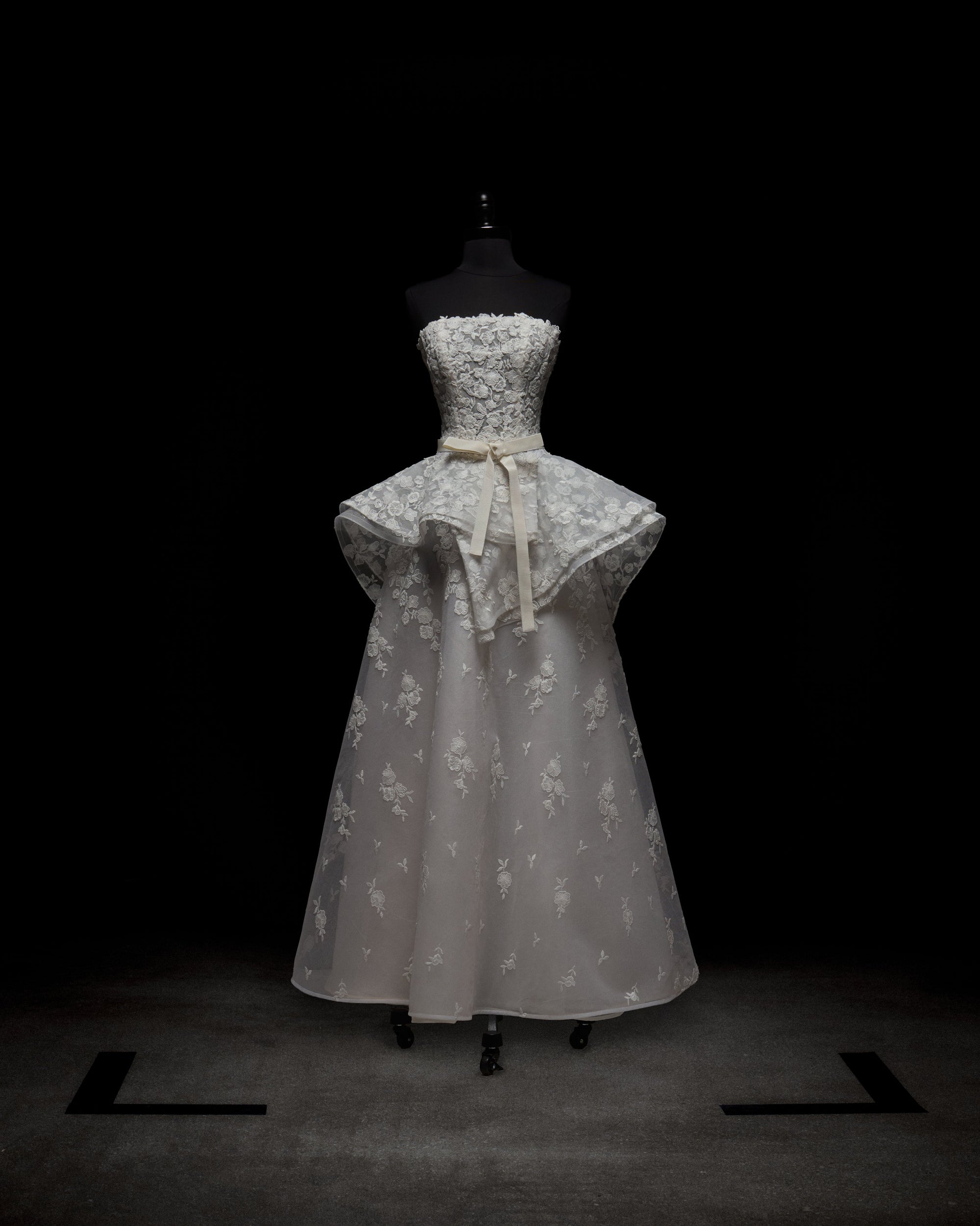 The Viviana Dress by SVT DESIGNS is a strapless white gown on display, featuring intricate lace detailing, a fitted bodice with a flared peplum, and a long flowing skirt adorned with floral patterns, complete with a waist-accentuating bow.