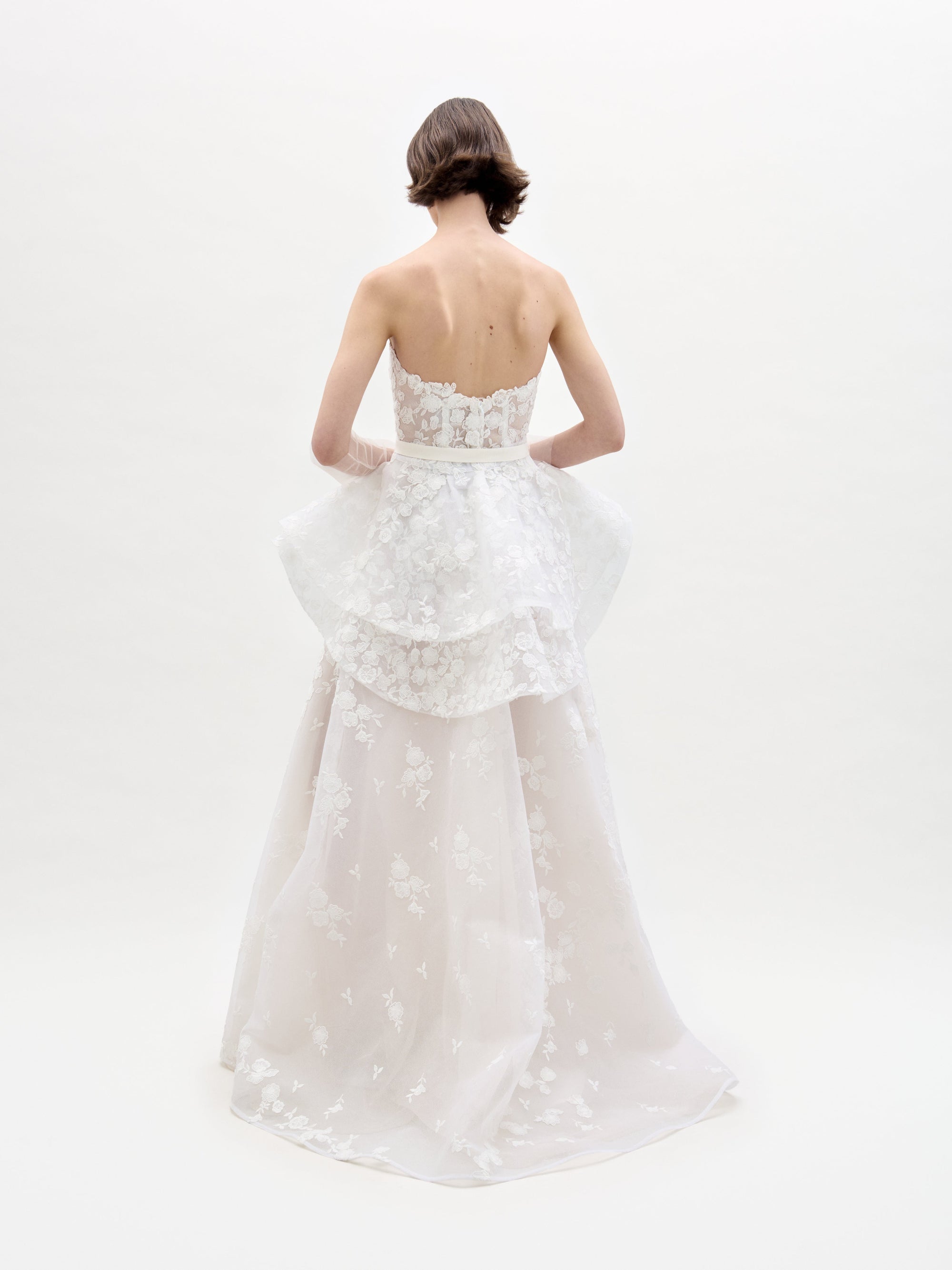 In a plain, bright setting, a person wears the strapless Viviana Dress by SVT DESIGNS. Their back faces the viewer, showcasing floral lace details with a layered peplum and sheer flowing skirt.