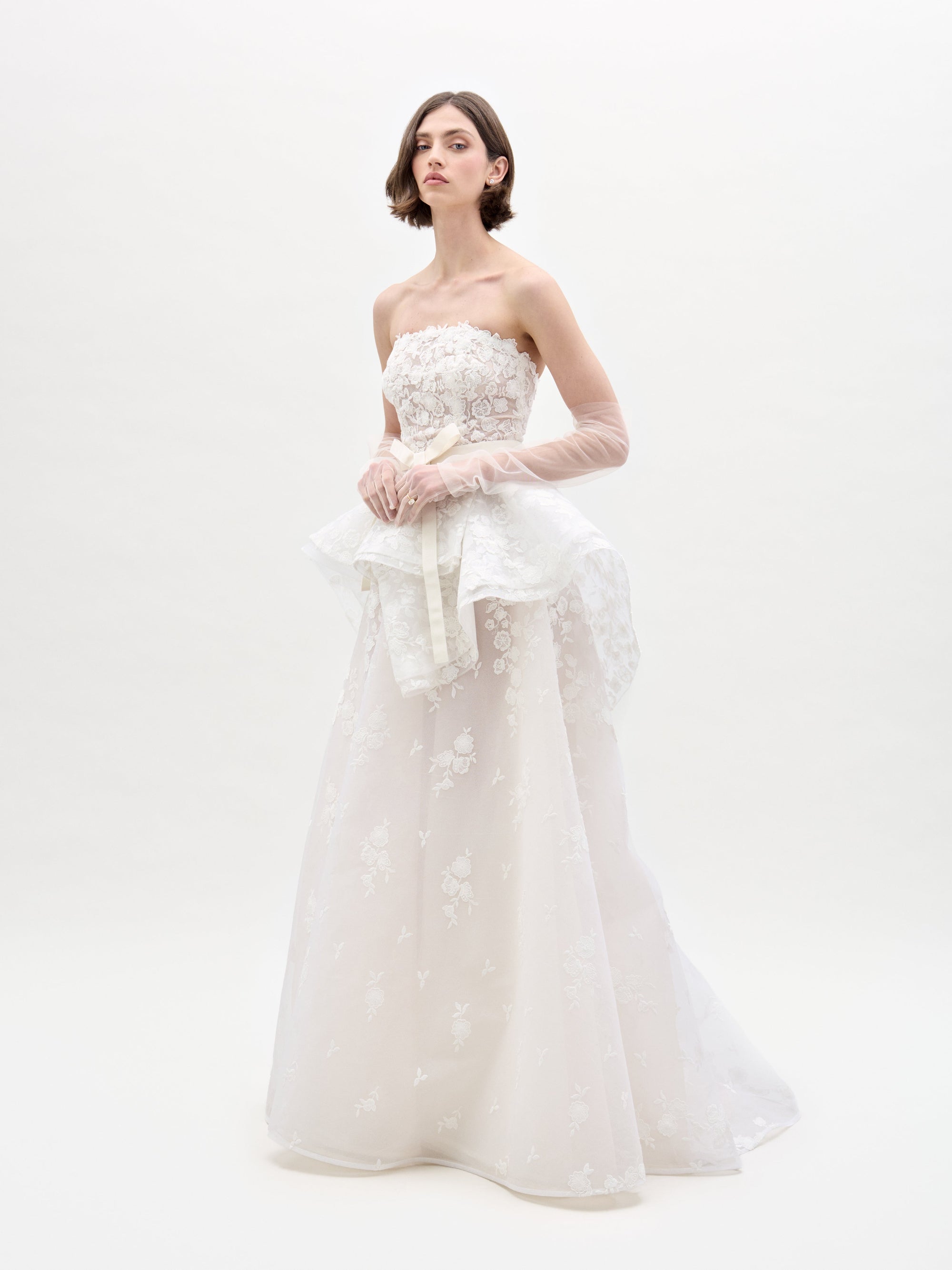 A person wears the Viviana Dress by SVT DESIGNS, a strapless white lace wedding gown adorned with floral patterns. The dress features a peplum waist and is paired with sheer lace gloves, all set against a plain white background.