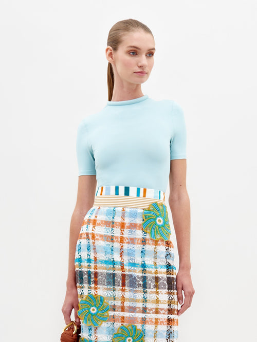 A woman dressed in a fitted Varinia Tshirt Turquoise and a multicolored plaid skirt stands against a plain white background, holding a small brown item in her right hand.