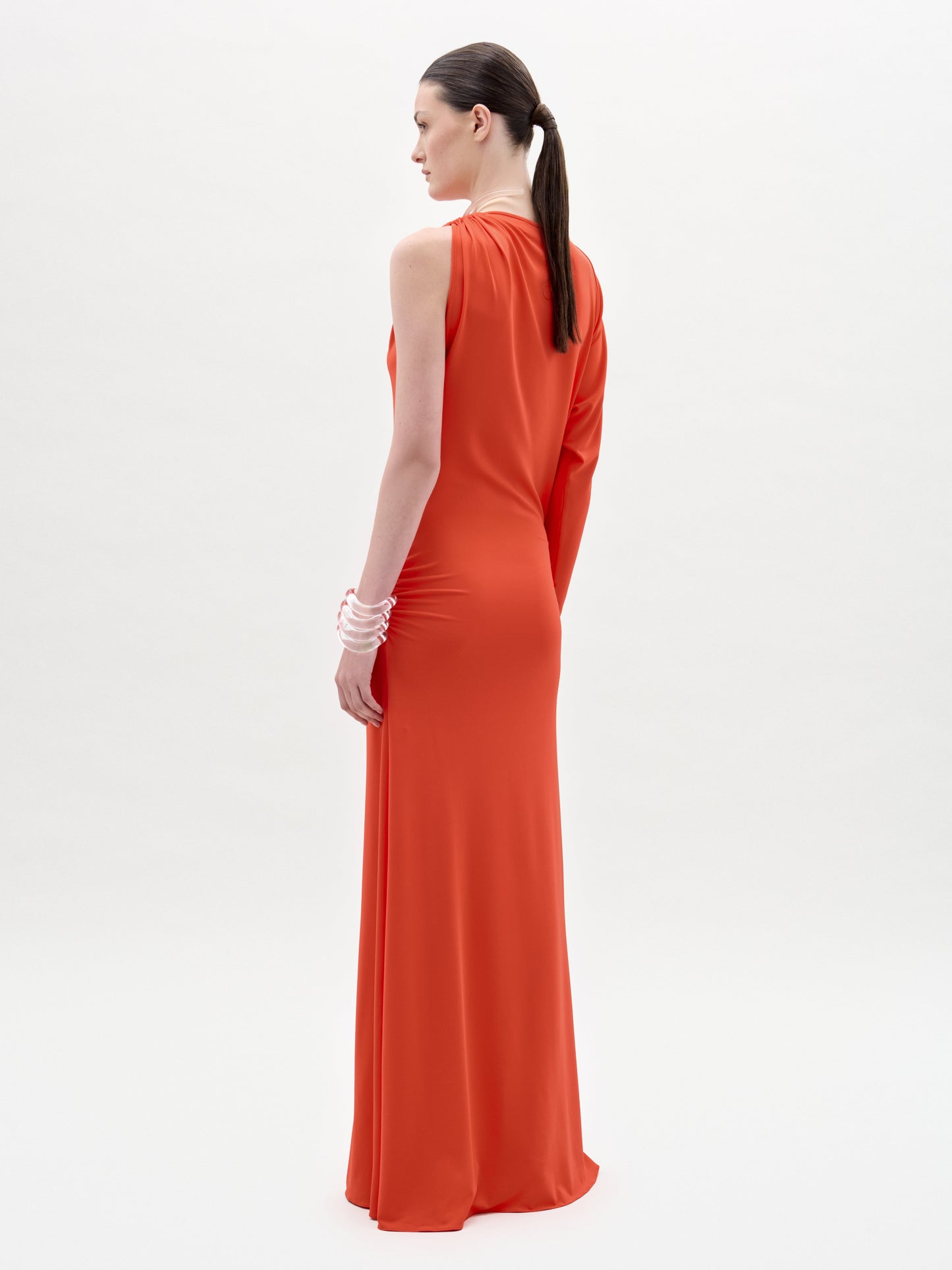 A woman faces away, wearing the SVT DESIGNS Virna Dress—a sleeveless orange maxi with a column silhouette in fine Italian fabric. Her hair is styled in a low ponytail and she wears white bangles, standing before a plain white background.