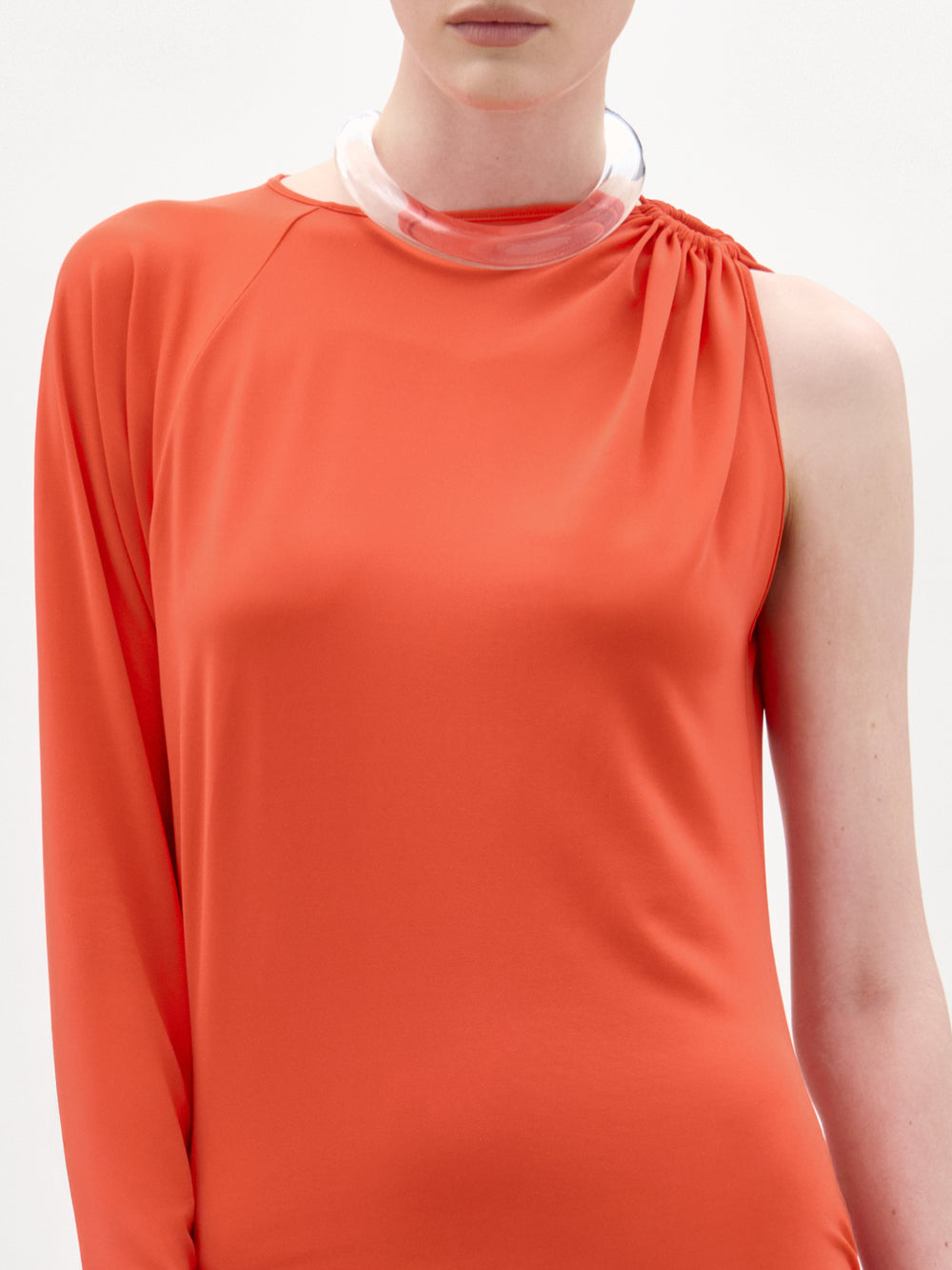 A person stands against a white background wearing SVT DESIGNS' Virna Dress—an asymmetrical orange Italian-fabric maxi with one long sleeve and one bare shoulder—accessorized with a clear chunky necklace.
