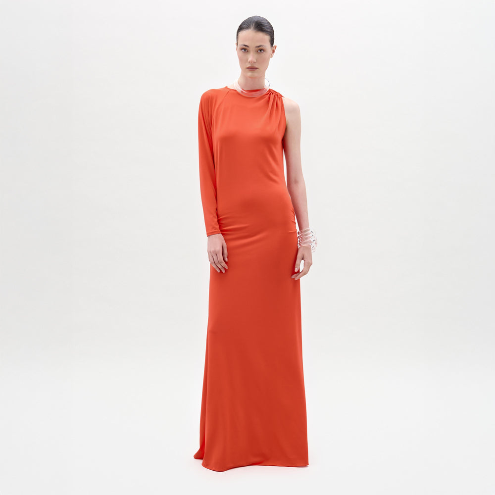 A woman stands against a white background wearing the SVT DESIGNS Virna Dress, a bright orange one-sleeve maxi with a column silhouette. Her hair is pulled back, styled with multiple bracelets and a necklace.