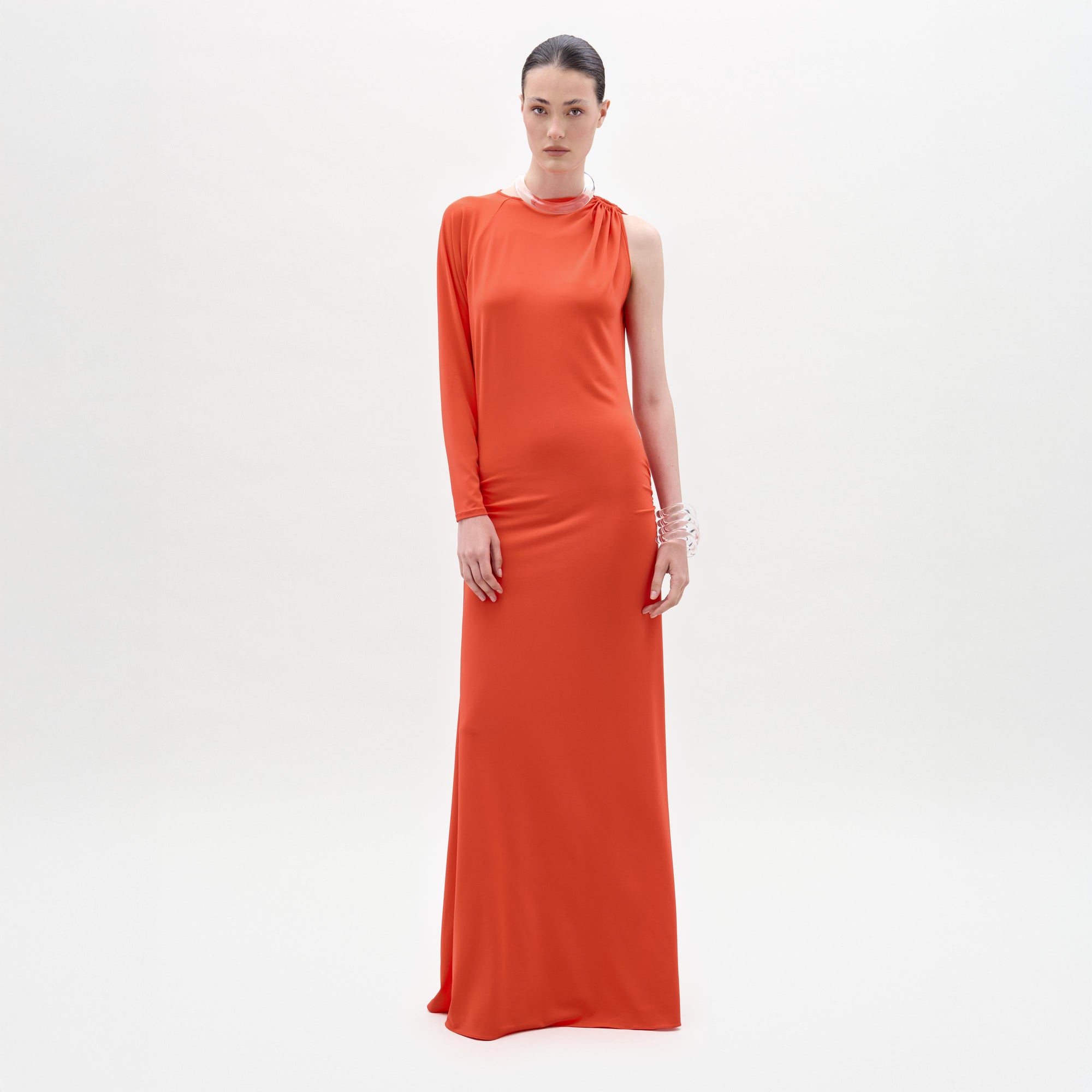 A woman stands against a white background wearing the SVT DESIGNS Virna Dress, a bright orange one-sleeve maxi with a column silhouette. Her hair is pulled back, styled with multiple bracelets and a necklace.