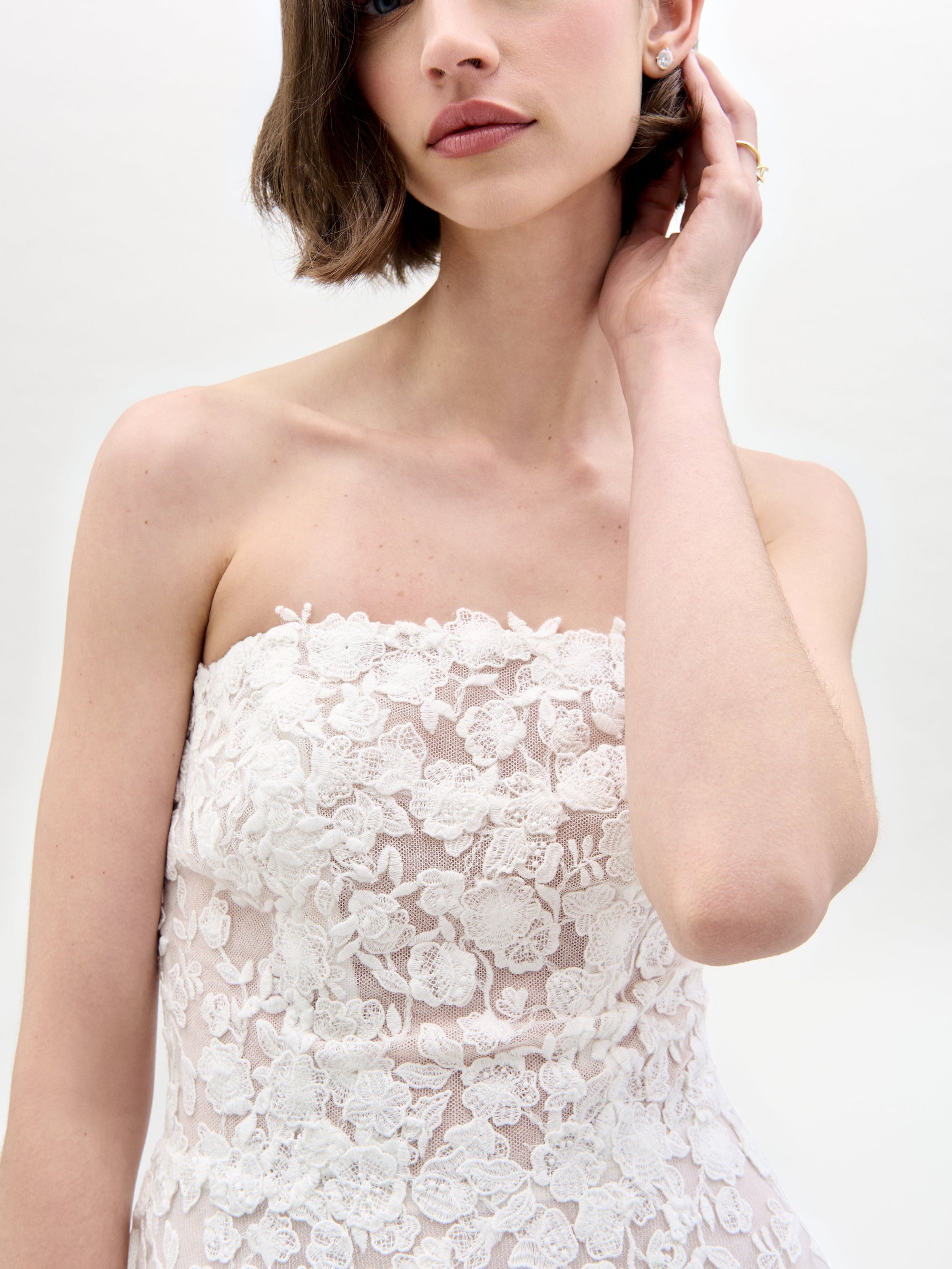 A woman with short brown hair wears the Viviana Dress—a strapless white lace dress made from luxurious Italian fabric—touching her neck. The photo, showing her upper torso and face, features a plain white background.