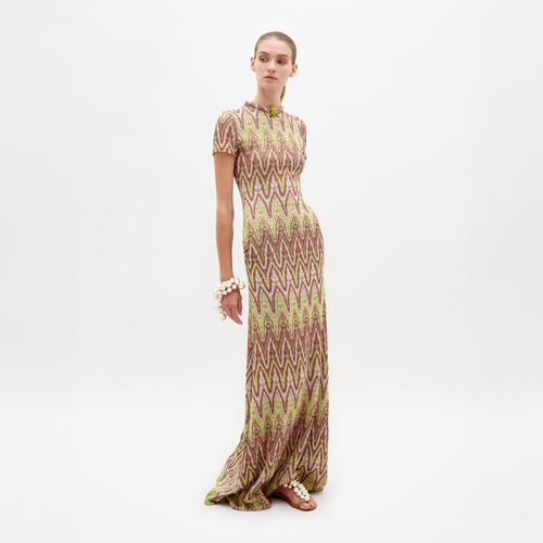 Wearing the Yara Dress by SVT DESIGNS, a person showcases its long, zigzag pattern in brown and light green inspired by Italian textile art. They have short hair, accessorized with large bracelets and sandals against a plain white backdrop.