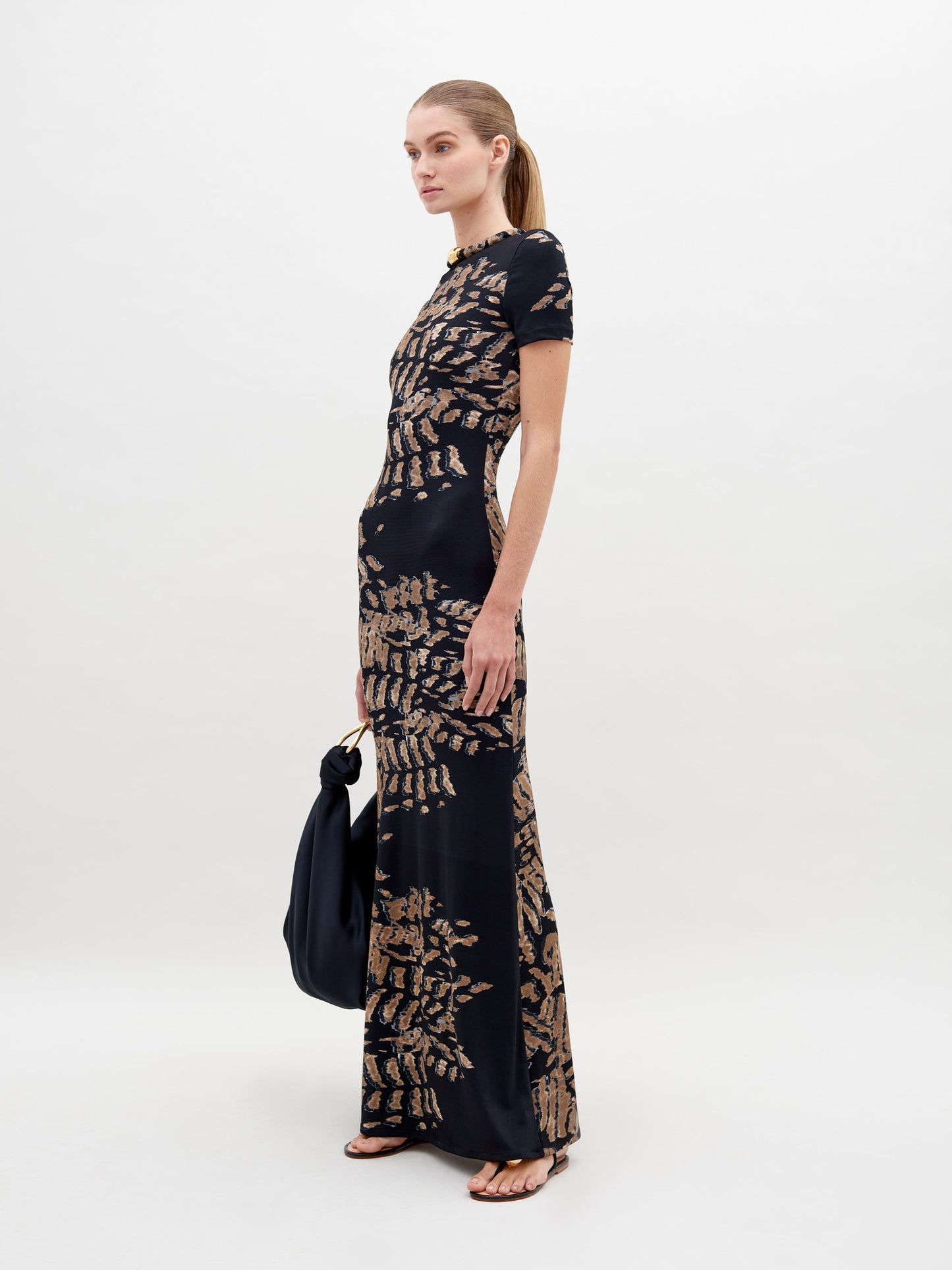 A woman stands against a plain white background wearing the Yara Dress Black Abstract Animal Print. She is holding a black bag and has her hair tied back in a low ponytail. Pre-order today for delivery by ship by date, November 15th, 2024.