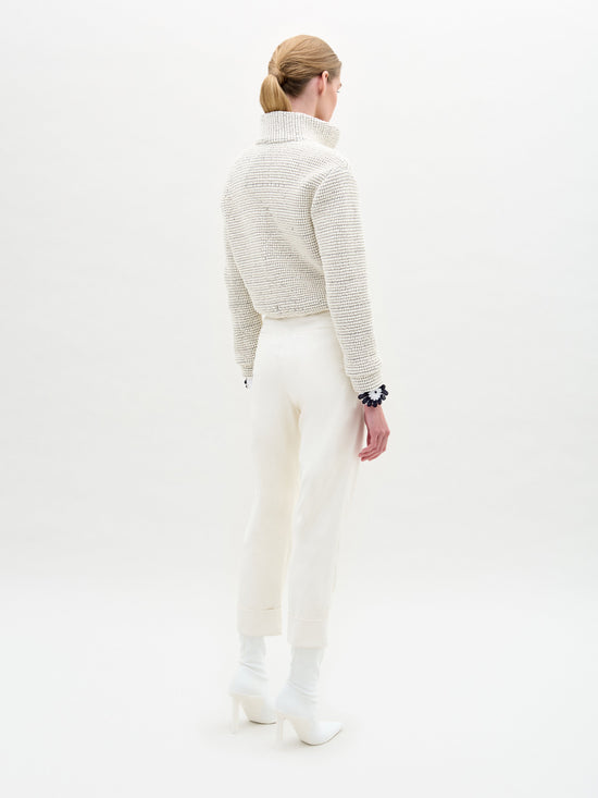 A blonde-haired person in a low ponytail faces away, wearing a textured cream turtleneck with SVT DESIGNS Moad Pant in Italian fabric and white high-waisted cropped style, paired with white heeled boots against a plain white background.