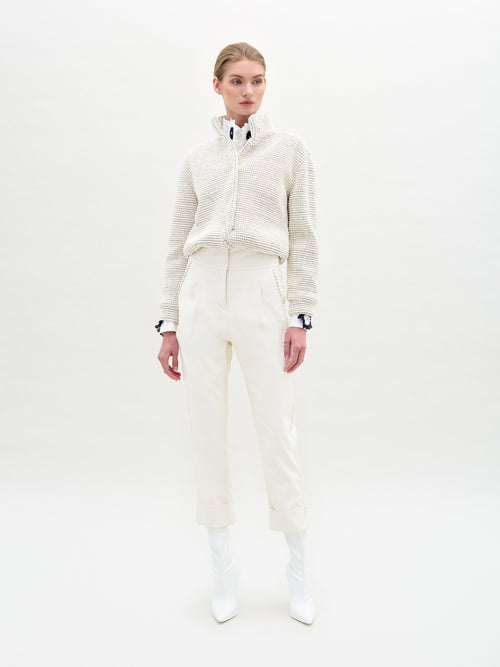 A person models the Moad Pant by SVT DESIGNS, showcasing a cream-colored textured jacket and an Italian fabric white collared shirt. Paired with high-waisted cream pants featuring a straight-leg fit, their look is completed by white boots against a plain white background.