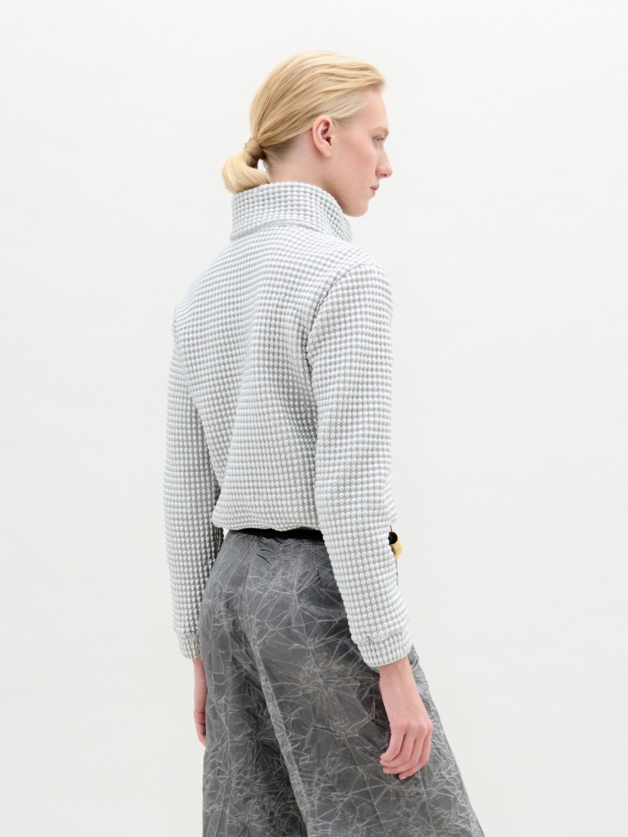 A person with blond hair tied back faces away from the camera against a light background, wearing the Zalta Jacket by SVT DESIGNS, a textured, oversized long-sleeve top paired with gray abstract-patterned trousers.
