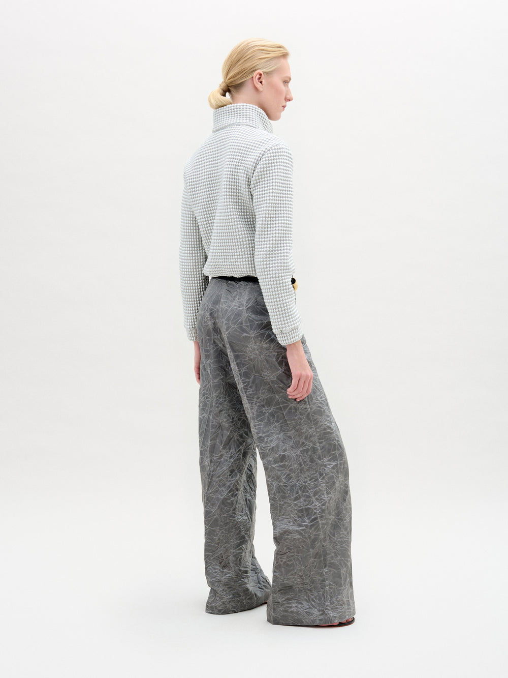 A person stands in a white textured zip-up jacket and gray Emine Pant with a gold belt against a plain white background.