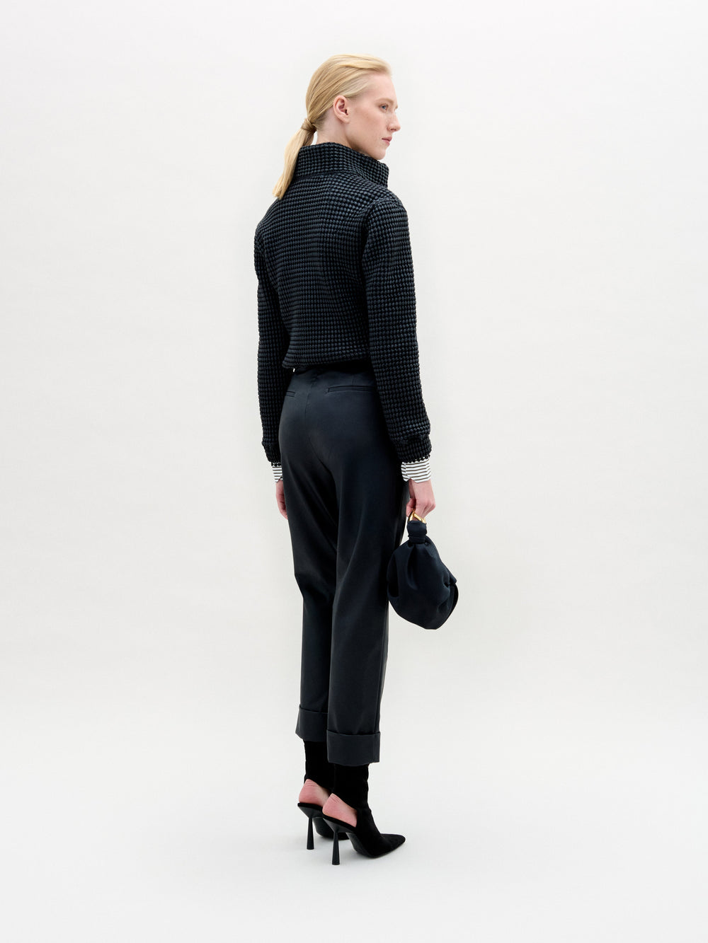 A person with light hair tied back faces away, wearing the high-collared Zalta Jacket by SVT DESIGNS in textured Italian fabric, dark cuffed pants, black heels, and holding a dark bag against a plain light background.