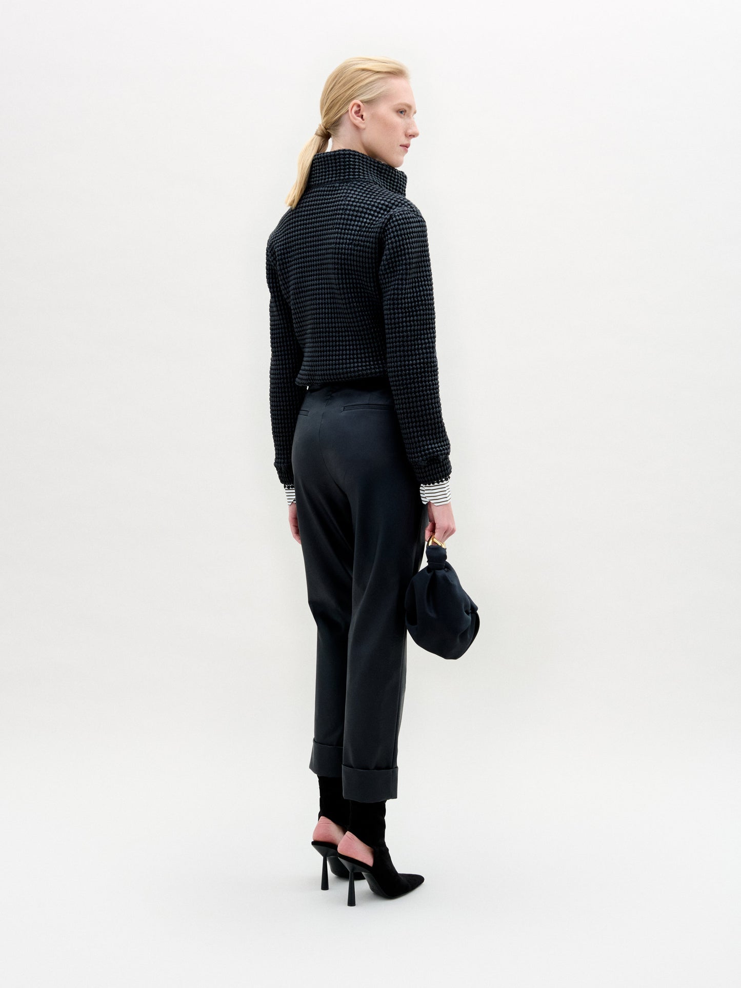 A person with light hair tied back faces away, wearing the high-collared Zalta Jacket by SVT DESIGNS in textured Italian fabric, dark cuffed pants, black heels, and holding a dark bag against a plain light background.