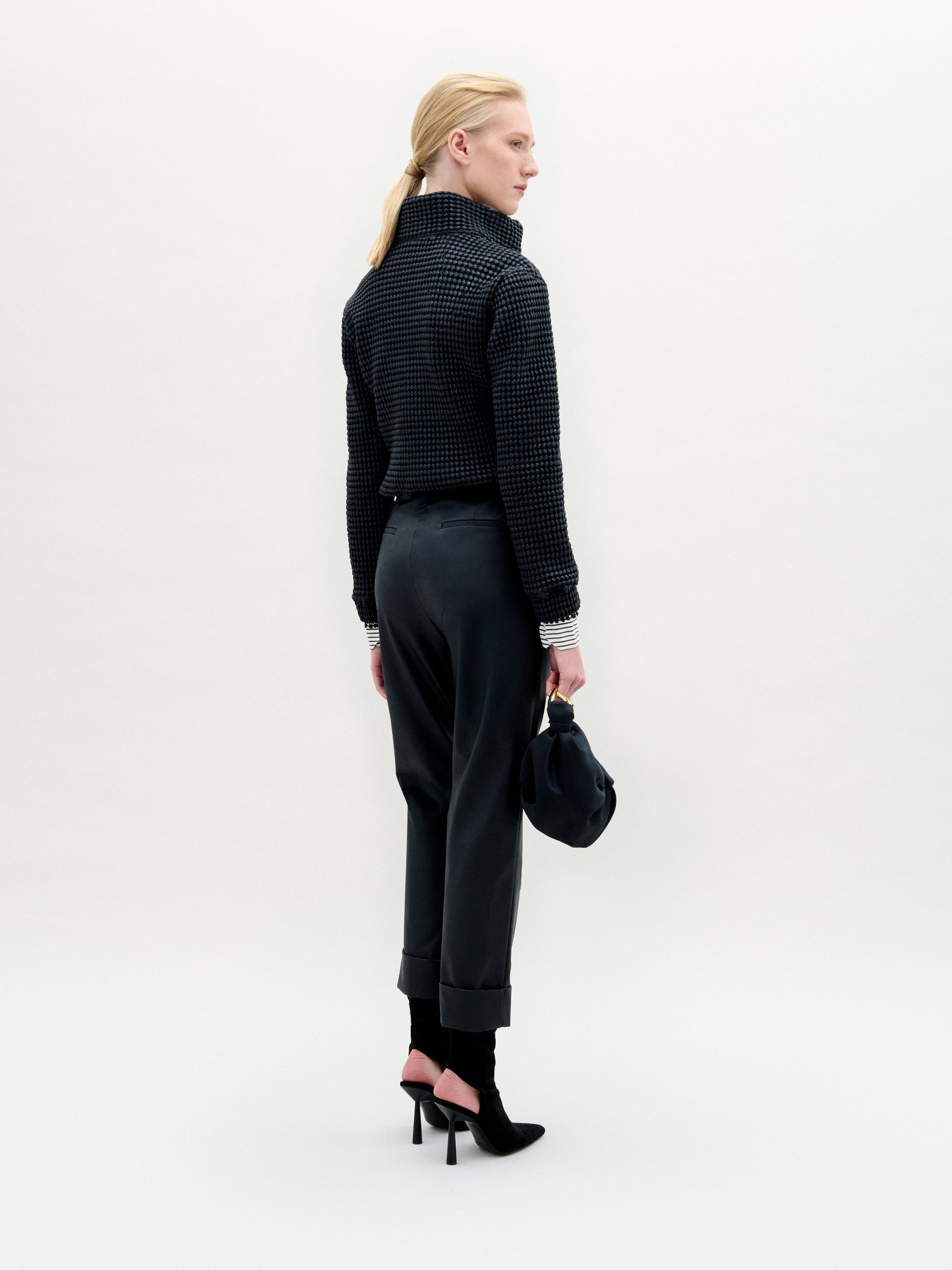 A person with light hair tied back faces away, wearing the high-collared Zalta Jacket by SVT DESIGNS in textured Italian fabric, dark cuffed pants, black heels, and holding a dark bag against a plain light background.