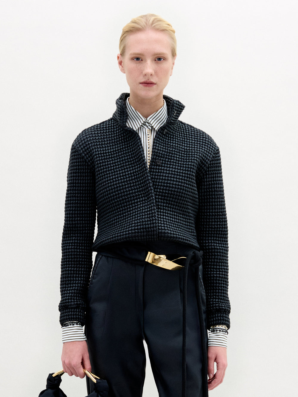 A person stands against a plain background wearing the Zalta Jacket by SVT DESIGNS over a striped button-up, dark high-waisted pants with a gold belt, and holds a bag with a knotted handle in their left hand.