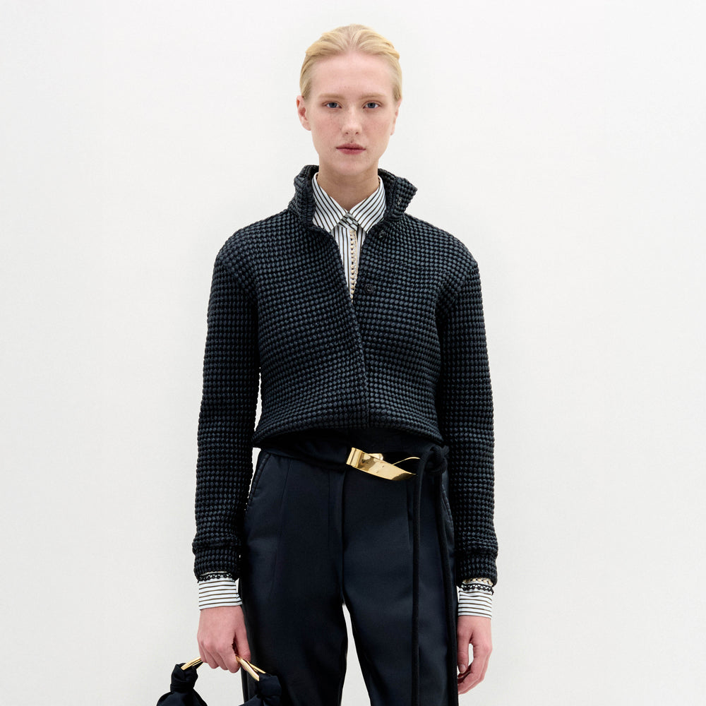 A person with short blond hair wears the Zalta Jacket by SVT DESIGNS—dark, cropped, and crafted from Italian fabric—over a striped collared shirt, dark high-waisted trousers with a gold belt, and holds a matching dark bag with gold accents against a plain white background.