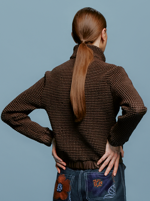 Wearing the Zalta Jacket Brown, a person with straight hair styled in a bun pairs it with blue pants adorned with decorative patches, poised elegantly against a blue background, ready for the spotlight on August 19th, 2024.