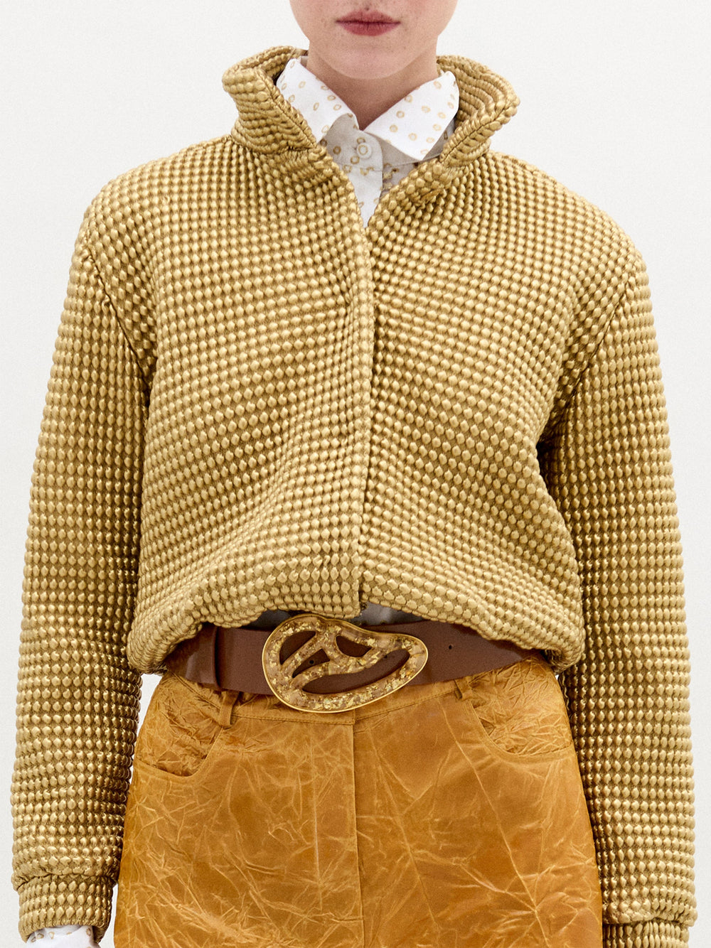 A blonde woman stands against a plain background wearing the Zalta jakcet in color Gold