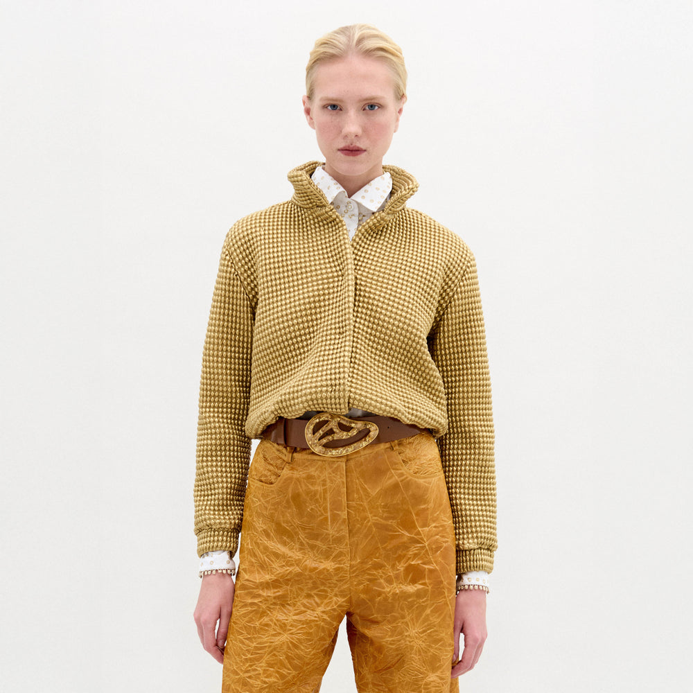 A person stands against a white background, wearing the SVT DESIGNS Zalta Jacket—an oversized yellow checkered cropped piece—styled over a white polka dot shirt with mustard textured pants and a brown belt featuring a large decorative buckle.
