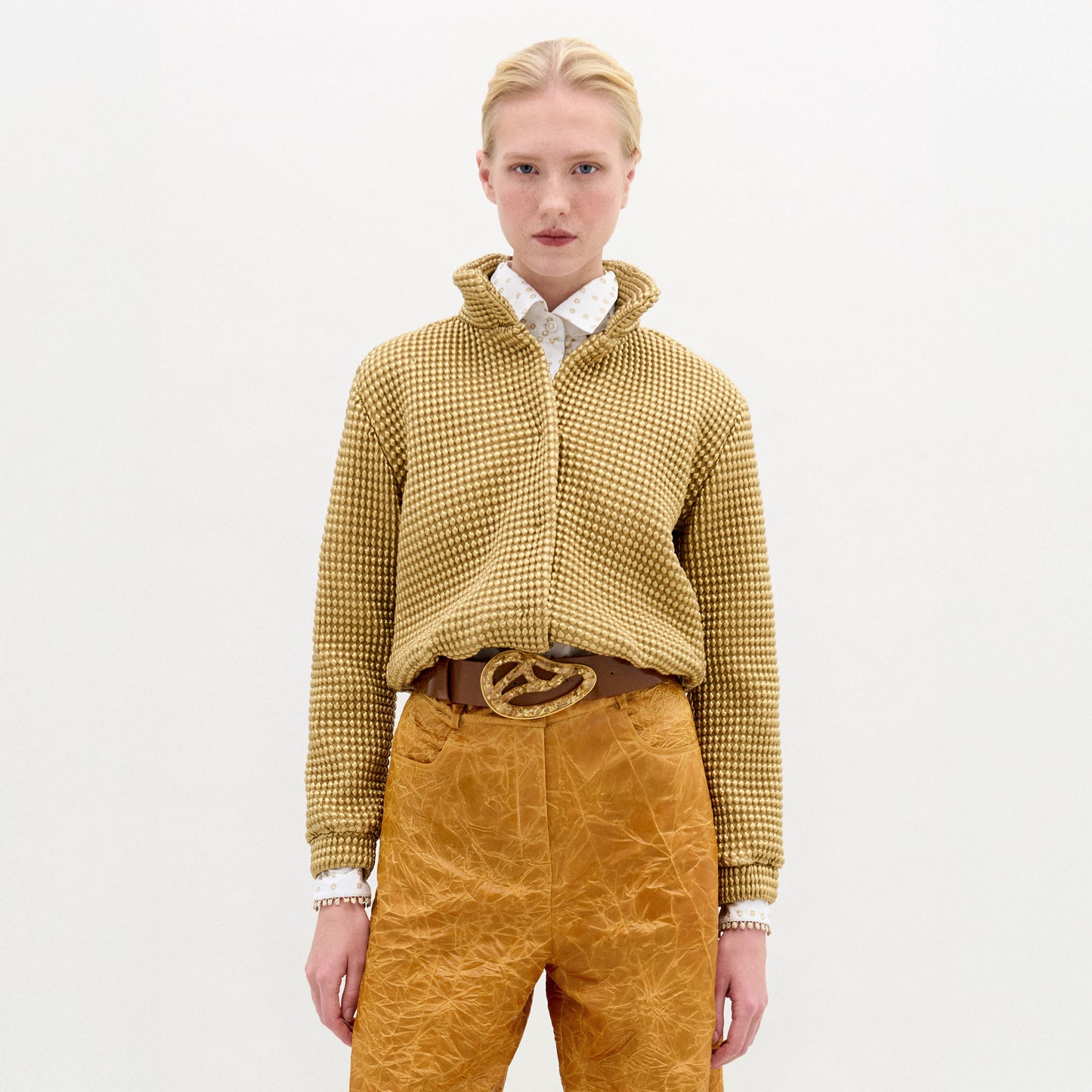 A person stands against a white background, wearing the SVT DESIGNS Zalta Jacket—an oversized yellow checkered cropped piece—styled over a white polka dot shirt with mustard textured pants and a brown belt featuring a large decorative buckle.