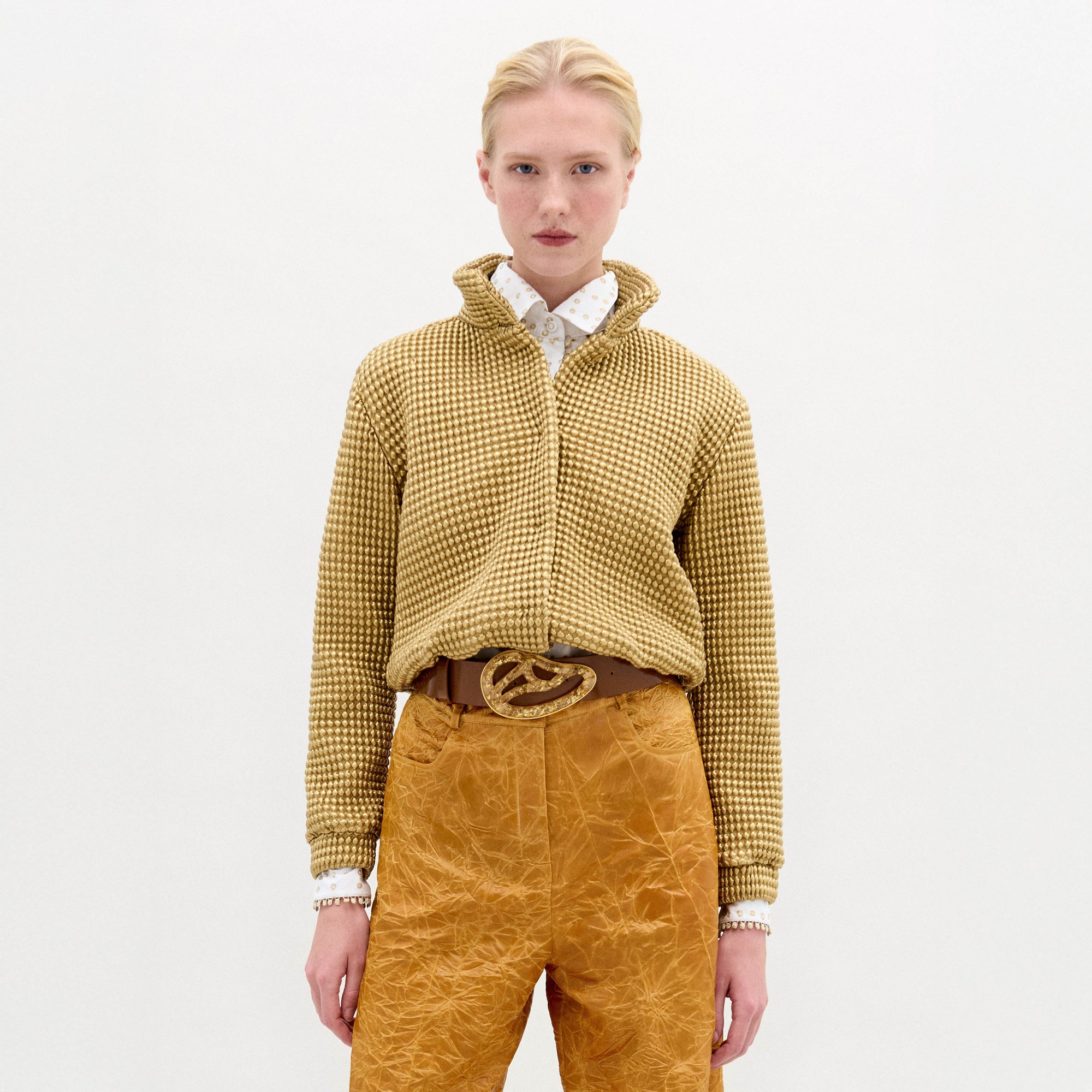 A blonde woman stands against a plain background wearing the Zalta jakcet in color Gold