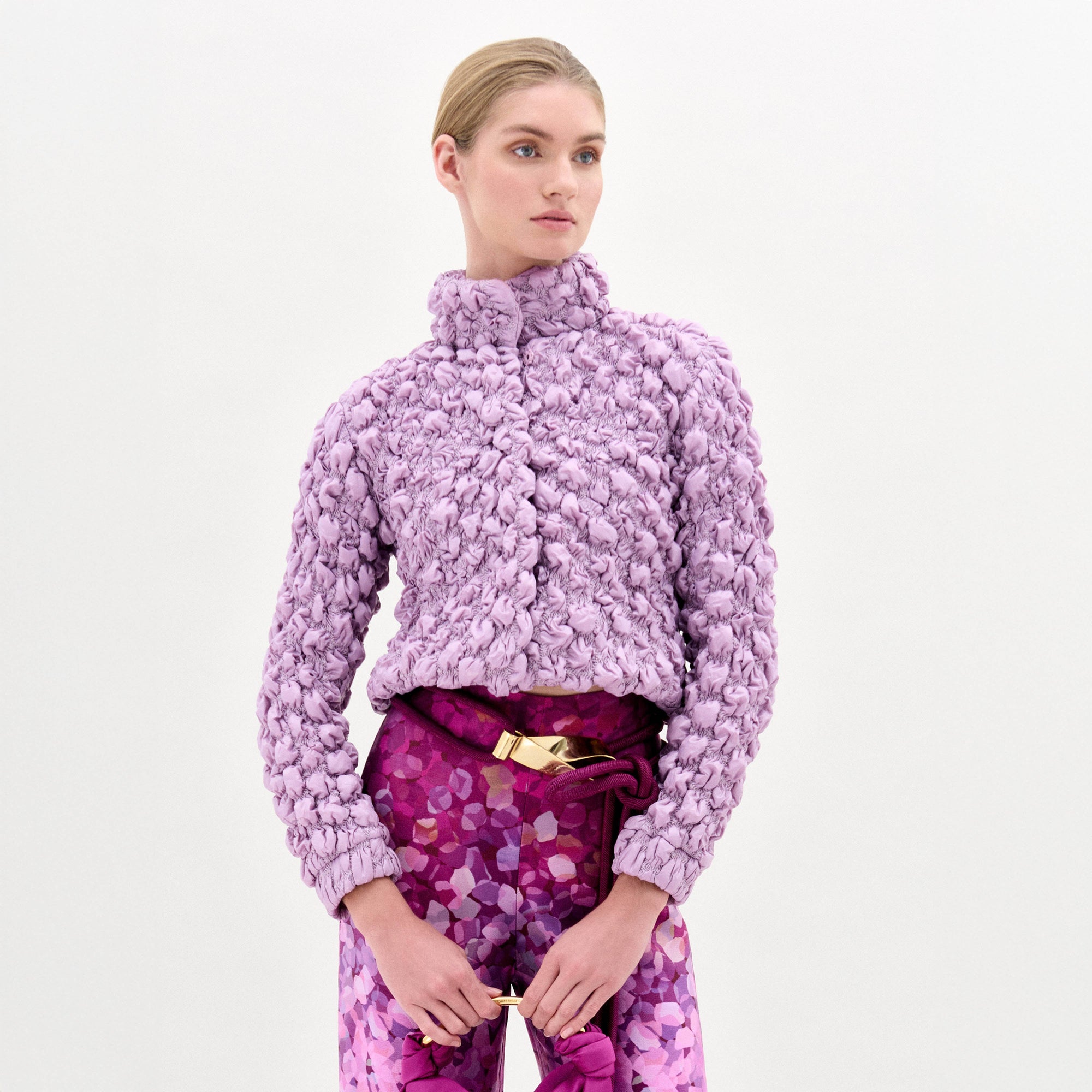 A woman poses against a white background wearing the oversized Zalta Jacket in textured lavender Italian fabric, paired with high-waisted pink patterned pants featuring a gold-buckled belt.