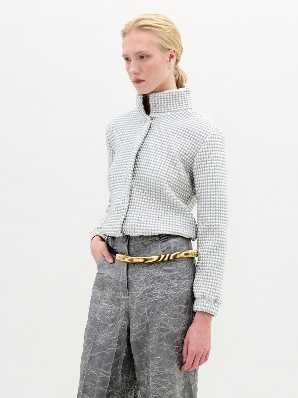 A person with light skin and blonde hair pulled back is wearing the Zalta Jacket by SVT DESIGNS, featuring a high collar and Italian fabric, paired with crinkled gray high-waisted pants and a beige rope-like belt against a white background.