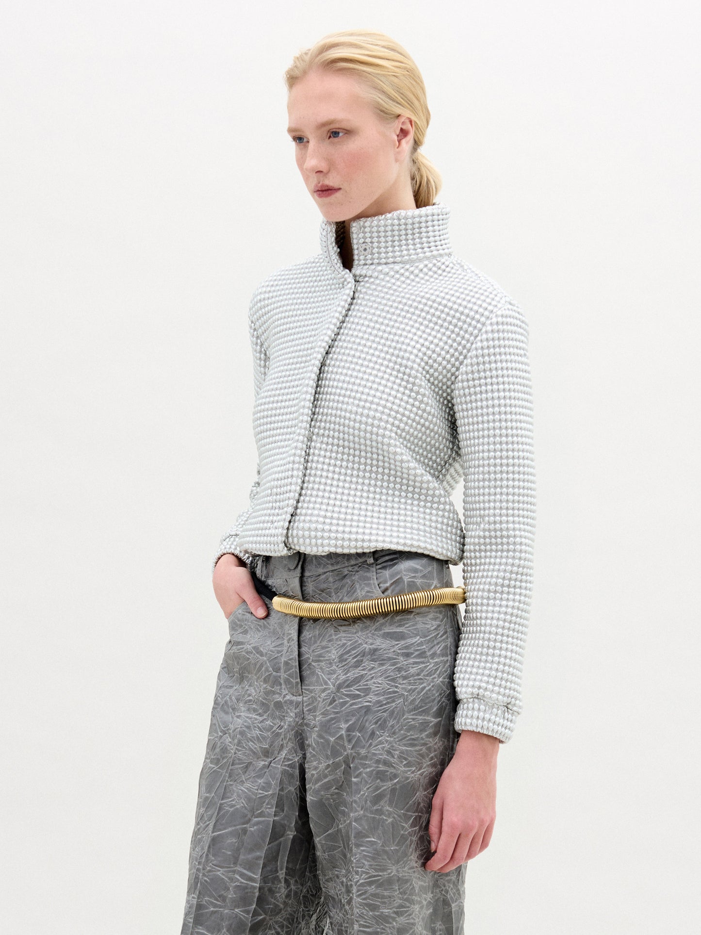 A person with light skin and blonde hair pulled back is wearing the Zalta Jacket by SVT DESIGNS, featuring a high collar and Italian fabric, paired with crinkled gray high-waisted pants and a beige rope-like belt against a white background.