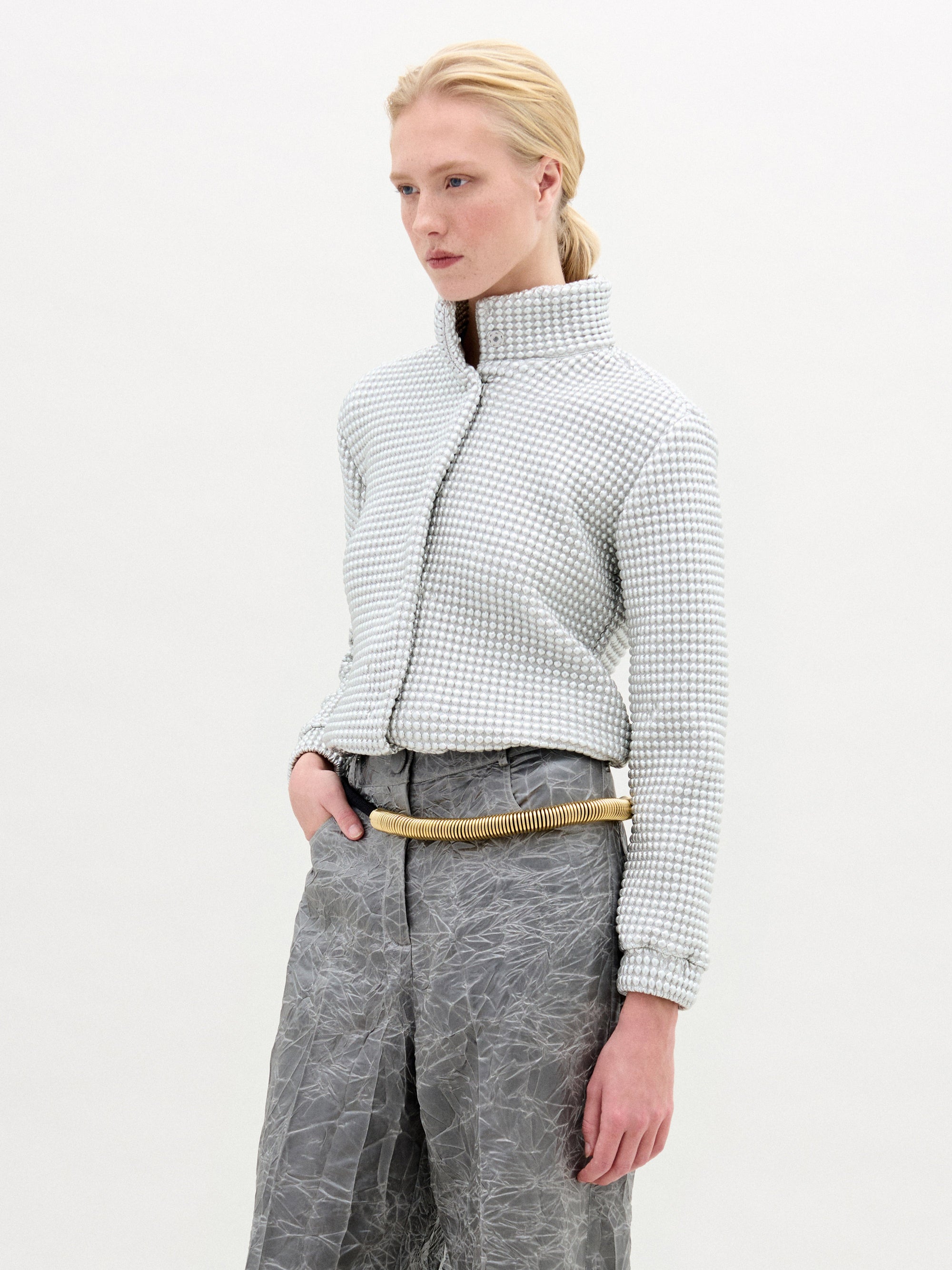 A person with light skin and blonde hair pulled back is wearing the Zalta Jacket by SVT DESIGNS, featuring a high collar and Italian fabric, paired with crinkled gray high-waisted pants and a beige rope-like belt against a white background.