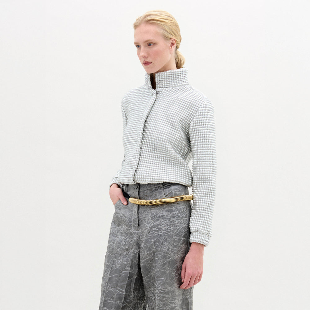 A person with light hair pulled back stands against a plain white background wearing the Zalta Jacket by SVT DESIGNS—a textured, light-colored, cropped Italian-fabric jacket with a high collar—paired with gray crinkled pants and hands in pockets.