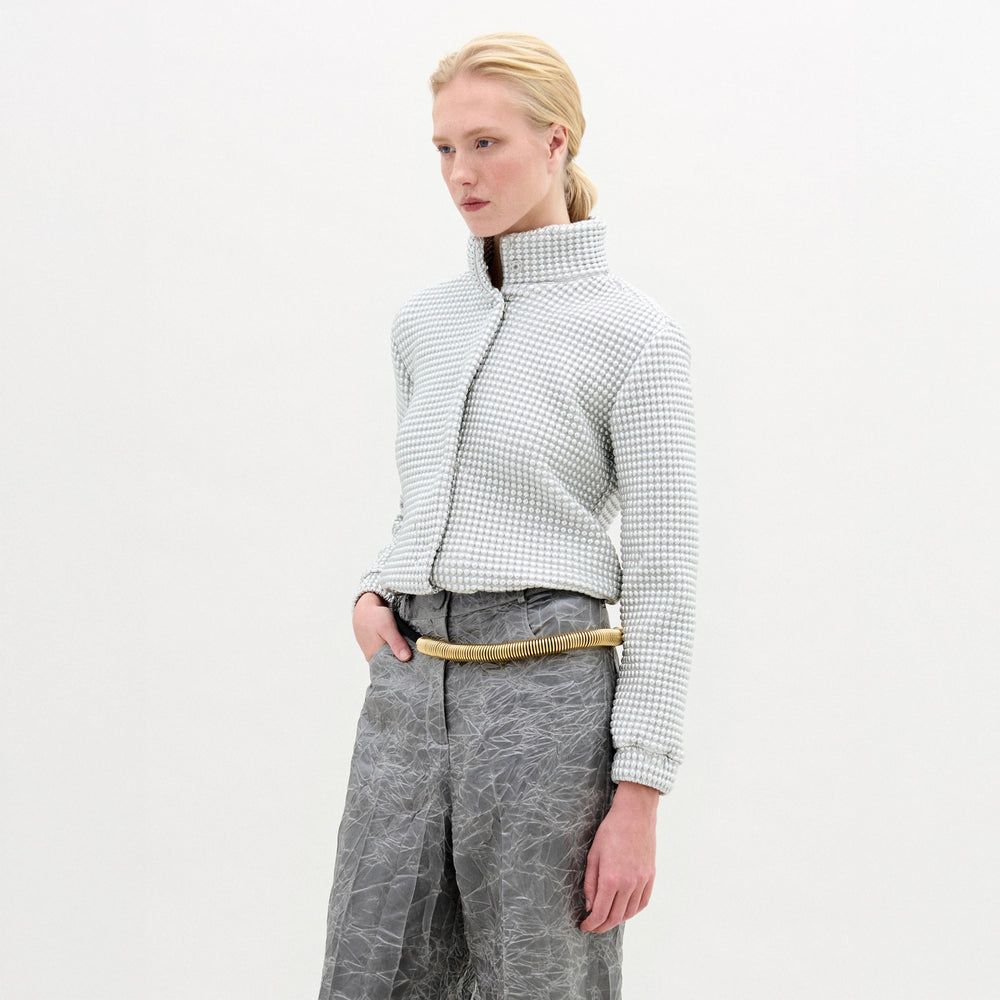 A person stands against a plain white background, wearing the oversized Zalta Jacket made from Italian fabric in a checkered pattern, paired with crinkled gray trousers and hands in pockets.