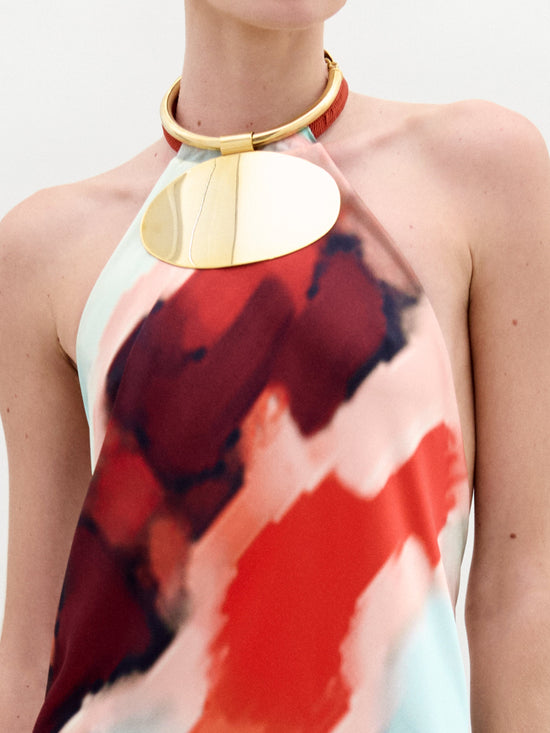 A person wears the SVT DESIGNS Zeffa Dress, a halter neck silk maxi with an abstract red, black, and white pattern. A gold circular pendant necklace decorates their neck. The backdrop is plain white with no visible face.