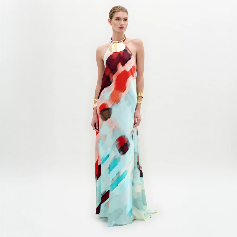 A woman poses against a white background in the SVT DESIGNS Zeffa Dress, a sleeveless, floor-length silk maxi featuring a bold abstract pattern in red, black, white, and aqua. She accessorizes with gold jewelry and wears her hair pulled back.