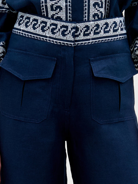 A person is wearing SVT DESIGNS' Zina Pant, crafted from Italian fabric in dark blue linen with large front pockets. Featuring intricate white geometric embroidery at the waistband and upper garment, the textured material completes this stylish look.