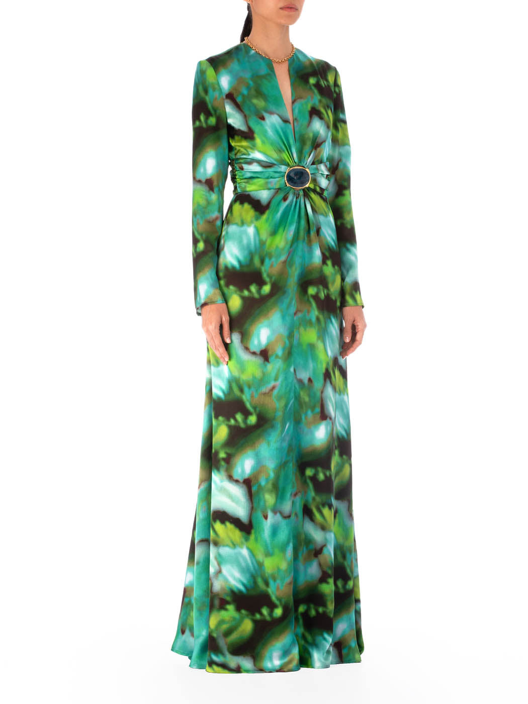 A person is wearing the Zarina Dress by SVT DESIGNS, a 100% silk maxi dress with long sleeves and a green and blue abstract multicolor print. It features a deep V-neck and a central circular embellishment at the waist.