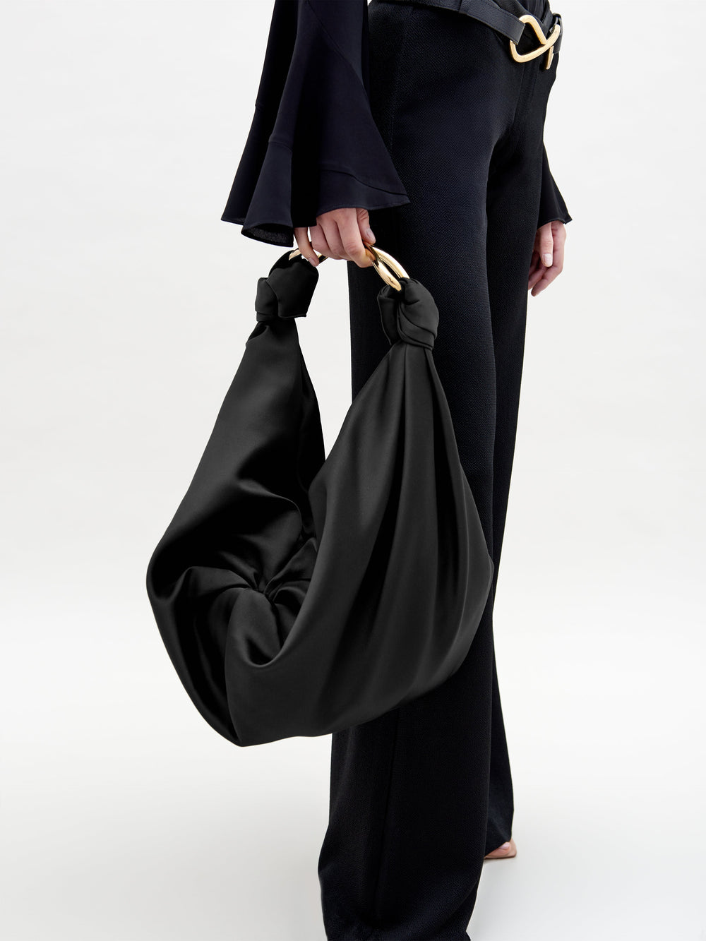 A person in black pants and a black top holds the Sofia Bag (Maxi), featuring gold handles and luxurious Italian textile, against a plain, light background that highlights the chic outfit and elegant accessory.