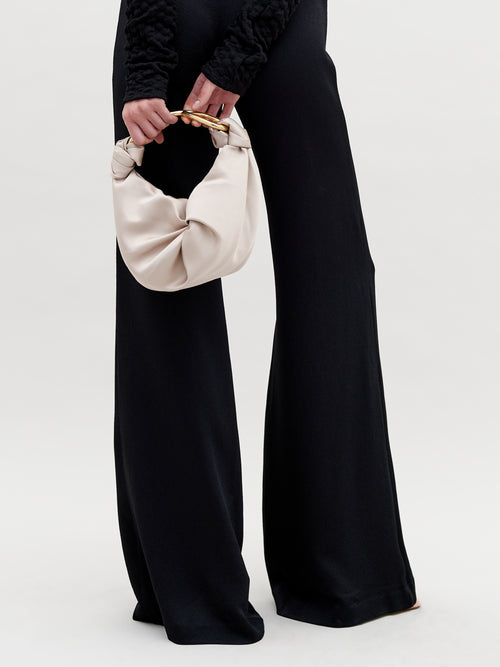 A person in black pants and a textured black top holds the Sofia Bag Small by SVT DESIGNS, featuring a light color and gold-toned circular handle, against a plain, light background.