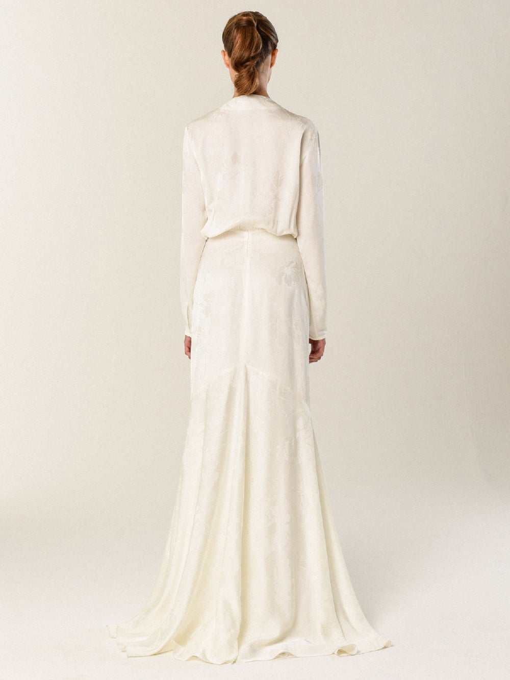 A woman faces away from the camera in SilviaTcherassi’s Albarella Dress—a long-sleeved, floor-length white jacquard gown with a subtle train and waist-defining bow, set against a plain, light background.