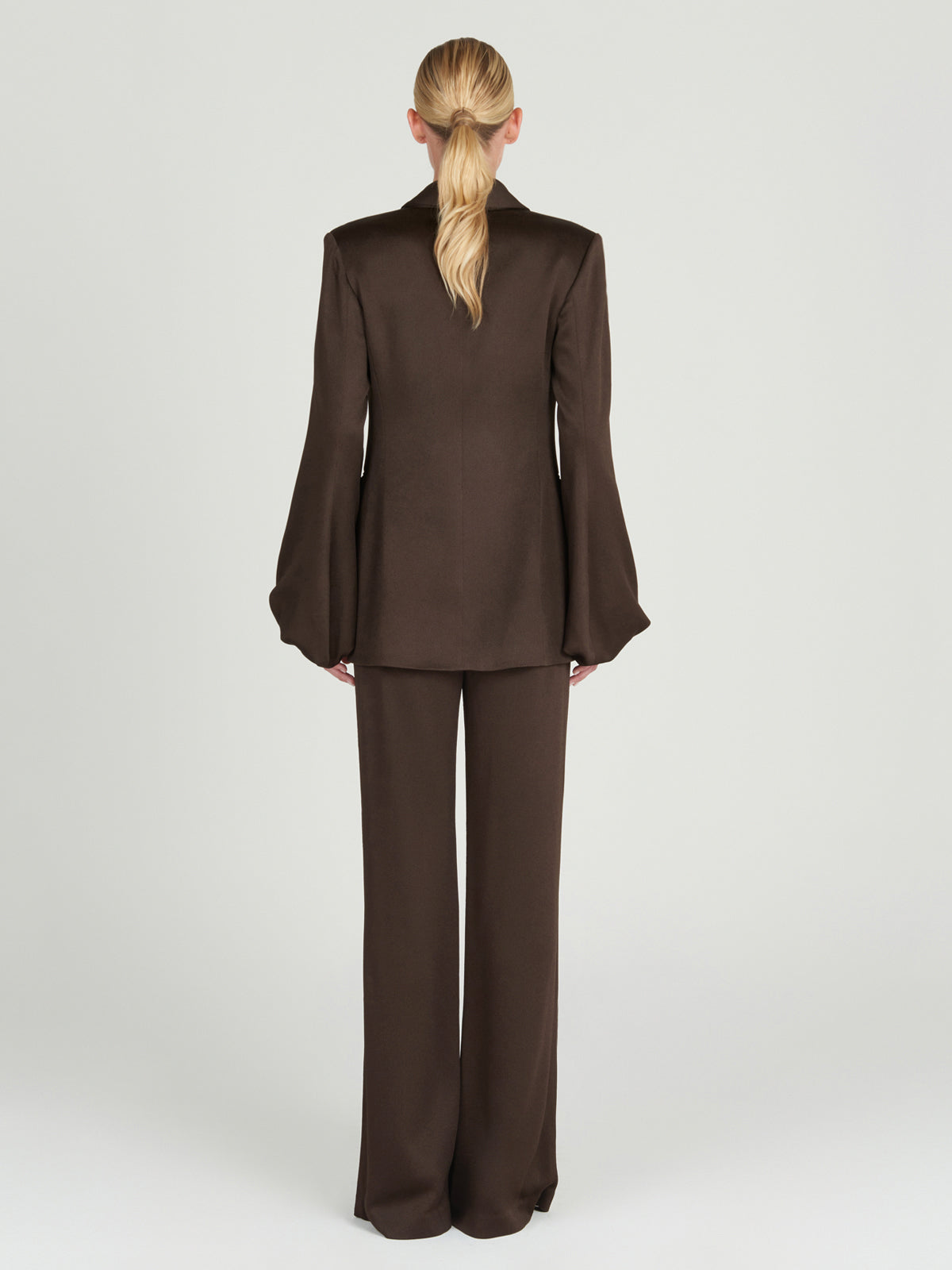 A person with long blonde hair tied back faces away from the camera, wearing Silvia Tcherassi's Palermo Pant—a high-waisted, wide-legged brown hammered satin suit—against a plain light background.