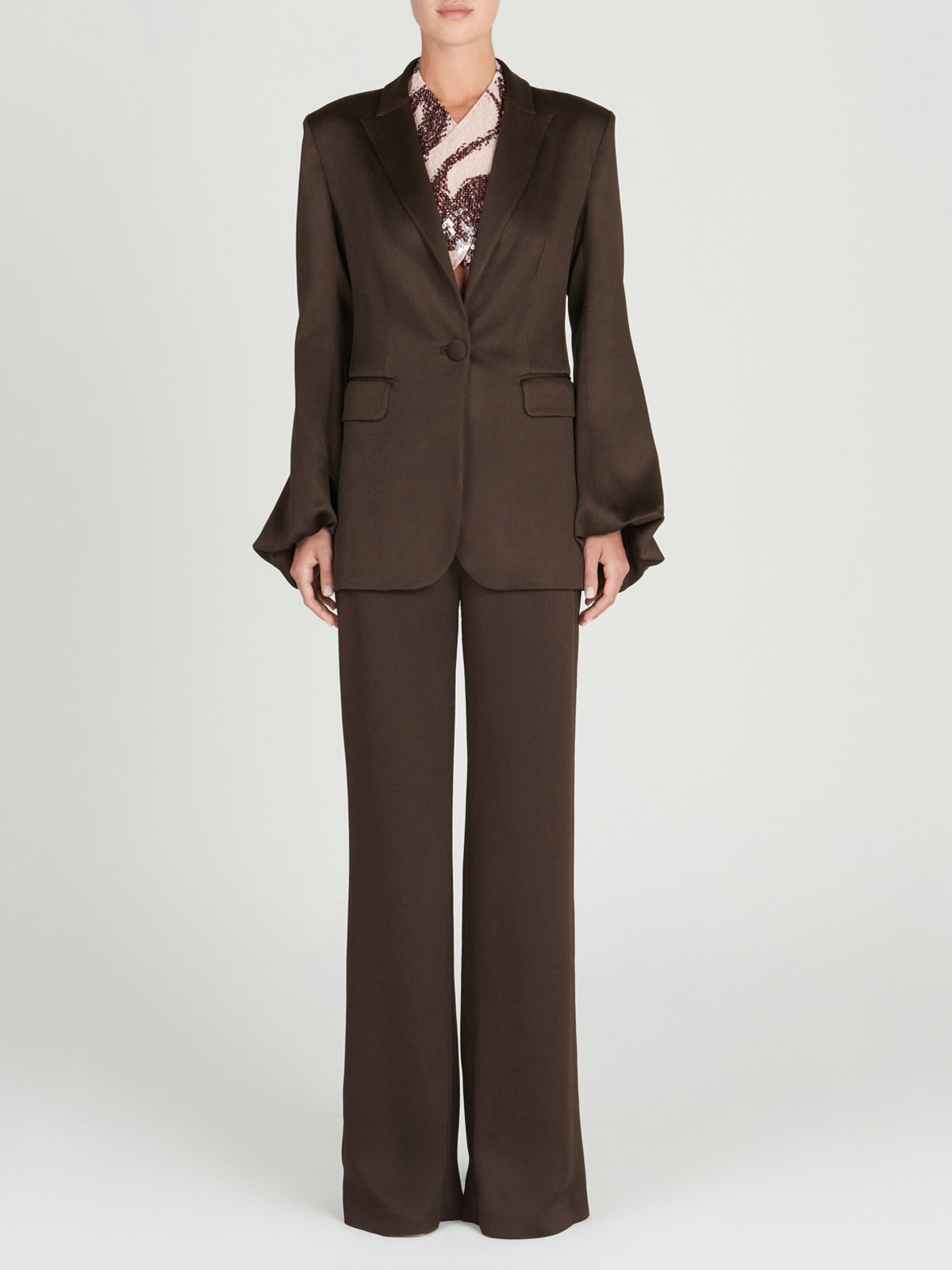 A person stands against a plain background wearing Silvia Tcherassi’s Palermo Pant, high-waisted with wide-legged flare, paired with a single-button blazer over a patterned blouse. Their arms are relaxed at their sides and their face is partially visible.