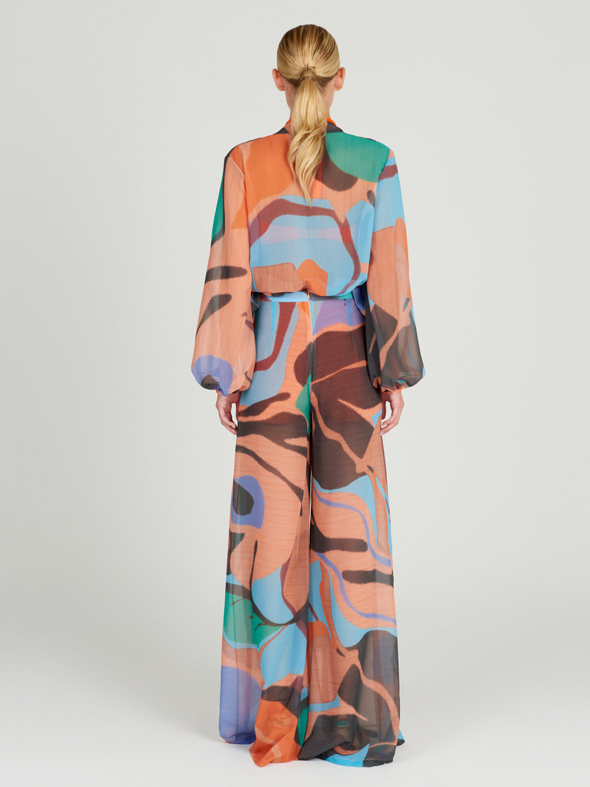 A person with blond hair in a low ponytail, seen from behind, wears Silvia Tcherassi's Benedetto Pant—a lightweight chiffon, wide-legged piece with abstract swirls and vibrant colors on a light background.