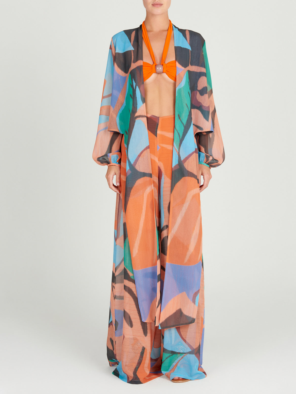 A person stands facing forward in the Silvia Tcherassi Benedetto Pant, crafted from lightweight chiffon with colorful abstract swirls, paired with a matching long-sleeve open jacket and an orange halter bikini top against a plain light background.