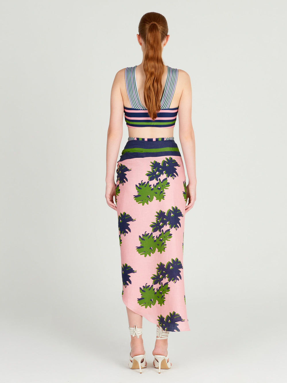 A woman with a ponytail faces away, wearing a striped crop top and the pink Mykonos Skirt by Silvia Tcherassi, featuring a green and navy blossom print, paired with white high-heeled sandals.