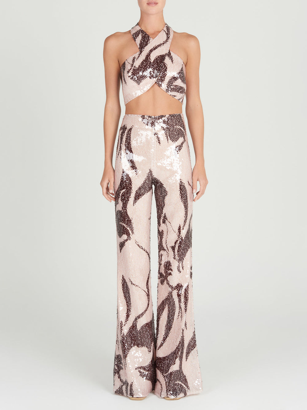 A person wearing the Nahia Top by Silvia Tcherassi, a shimmering two-piece with a crisscross halter neckline and high-waisted wide-leg pants. The metallic-toned abstract pattern stands out against a light background, and their hands are by their sides.