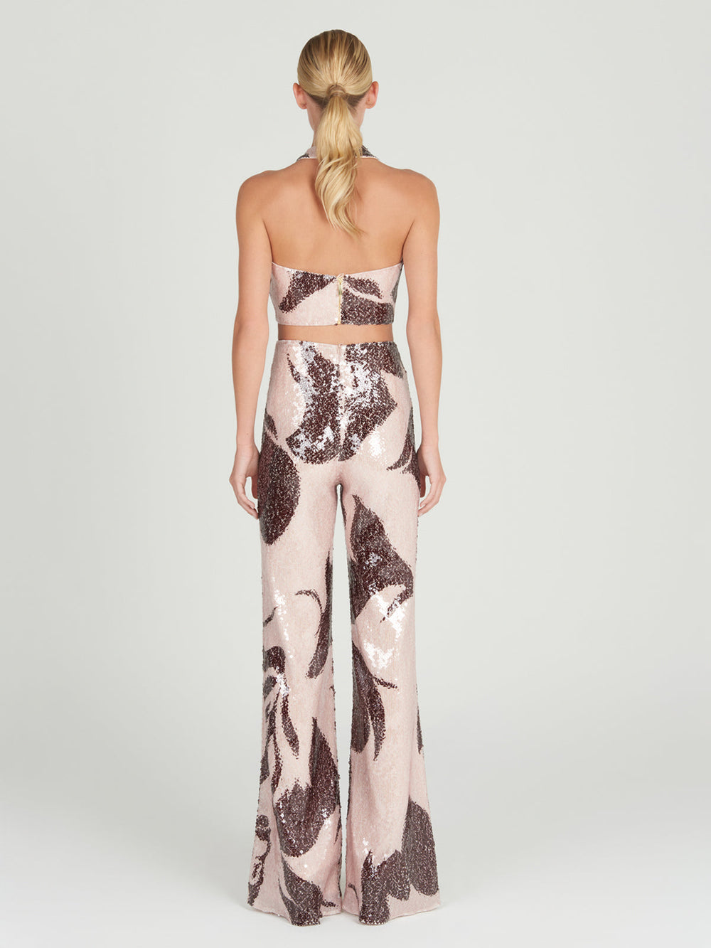 A person with long blonde hair faces away, wearing the strapless, backless Nahia Top by Silvia Tcherassi paired with flared pants. Both items shimmer with large brown and pink sequins under bright lighting. The stylish outfit includes a subtle gold zipper against a plain white backdrop.
