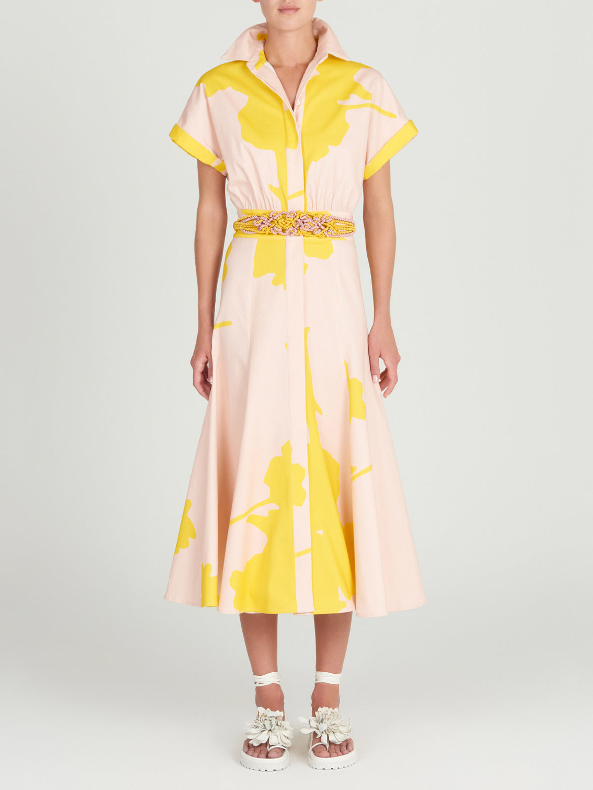 A person models the Noor Dress by Silvia Tcherassi, featuring a flattering cut with large yellow florals on a pink knee-length canvas, short sleeves, and a classic shirt collar, paired with a colorful braided belt and white sandals with flower details.