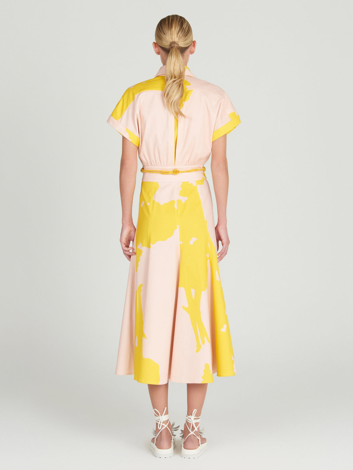 A person with long blonde hair stands facing away, wearing a light pink Noor Dress by Silvia Tcherassi. The knee-length dress features bold yellow abstract patterns, a flattering cut, and a classic shirt collar. It's paired with white lace-up shoes against a plain, light-colored background.