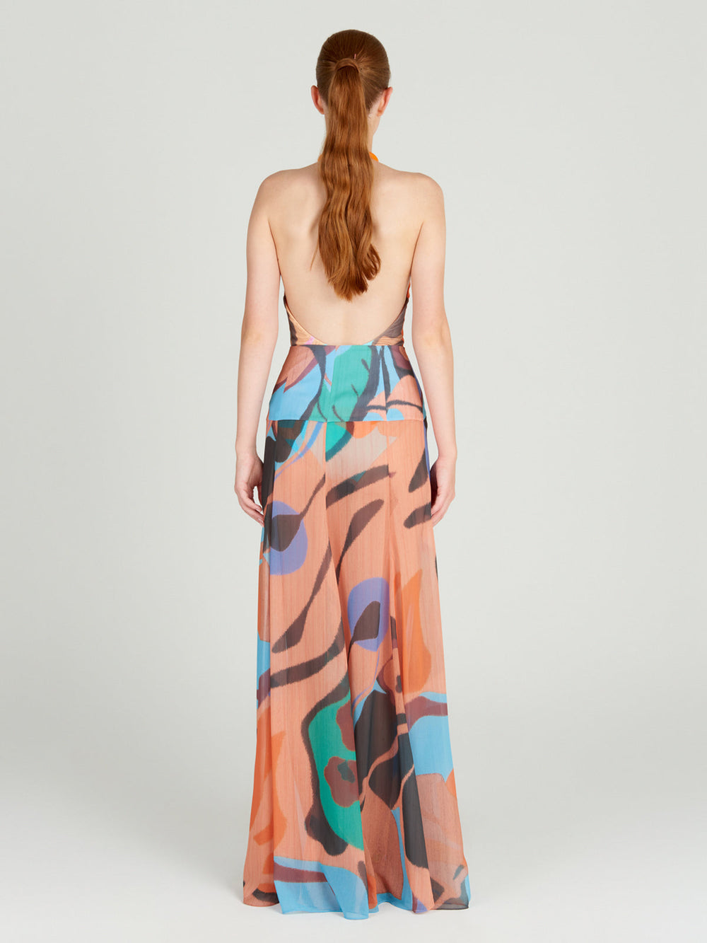 A person with long red hair stands facing away in a backless maxi dress crafted from lightweight chiffon, featuring a colorful abstract pattern of orange, blue, green, and black on a light backdrop. The setting is plain and neutral. Skirt by Silvia Tcherassi called Cagliari.