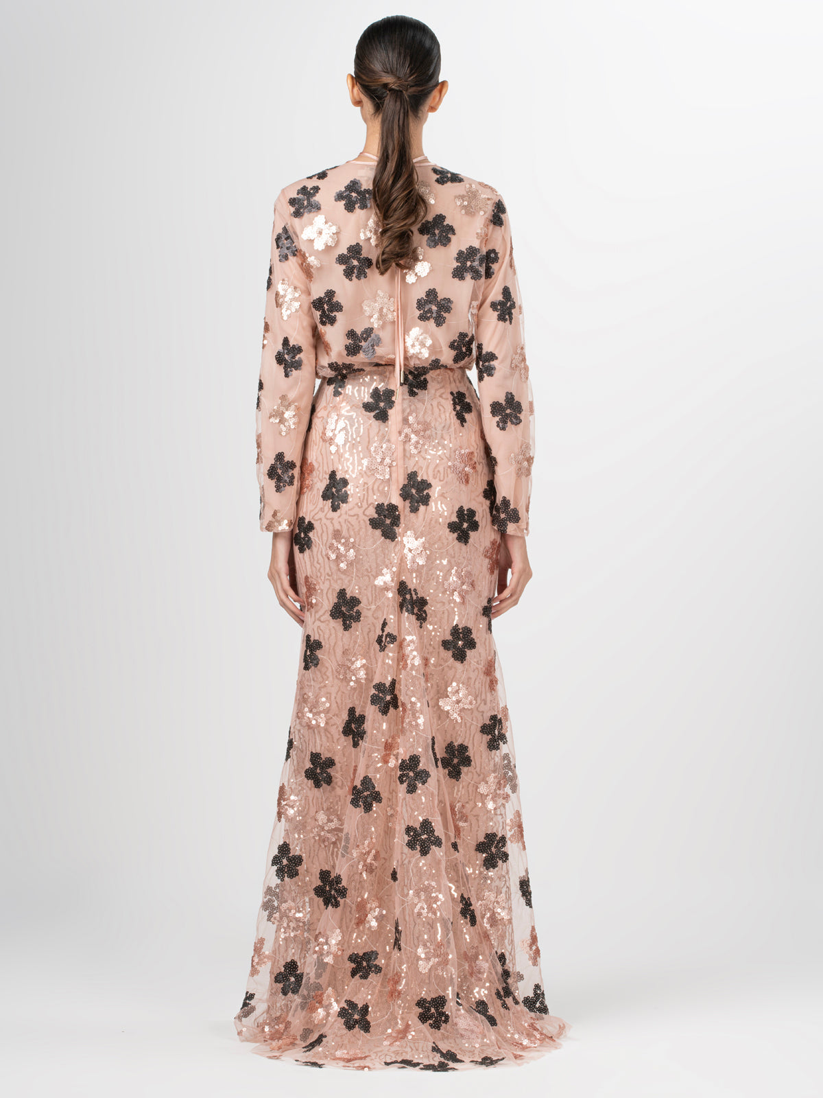 A person with long hair is shown from the back wearing the Tosca Blouse by Silvia Tcherassi US, a sheer full-length gown embellished with black floral sequins lace over a beige underlayer, featuring long sleeves against a plain white background.